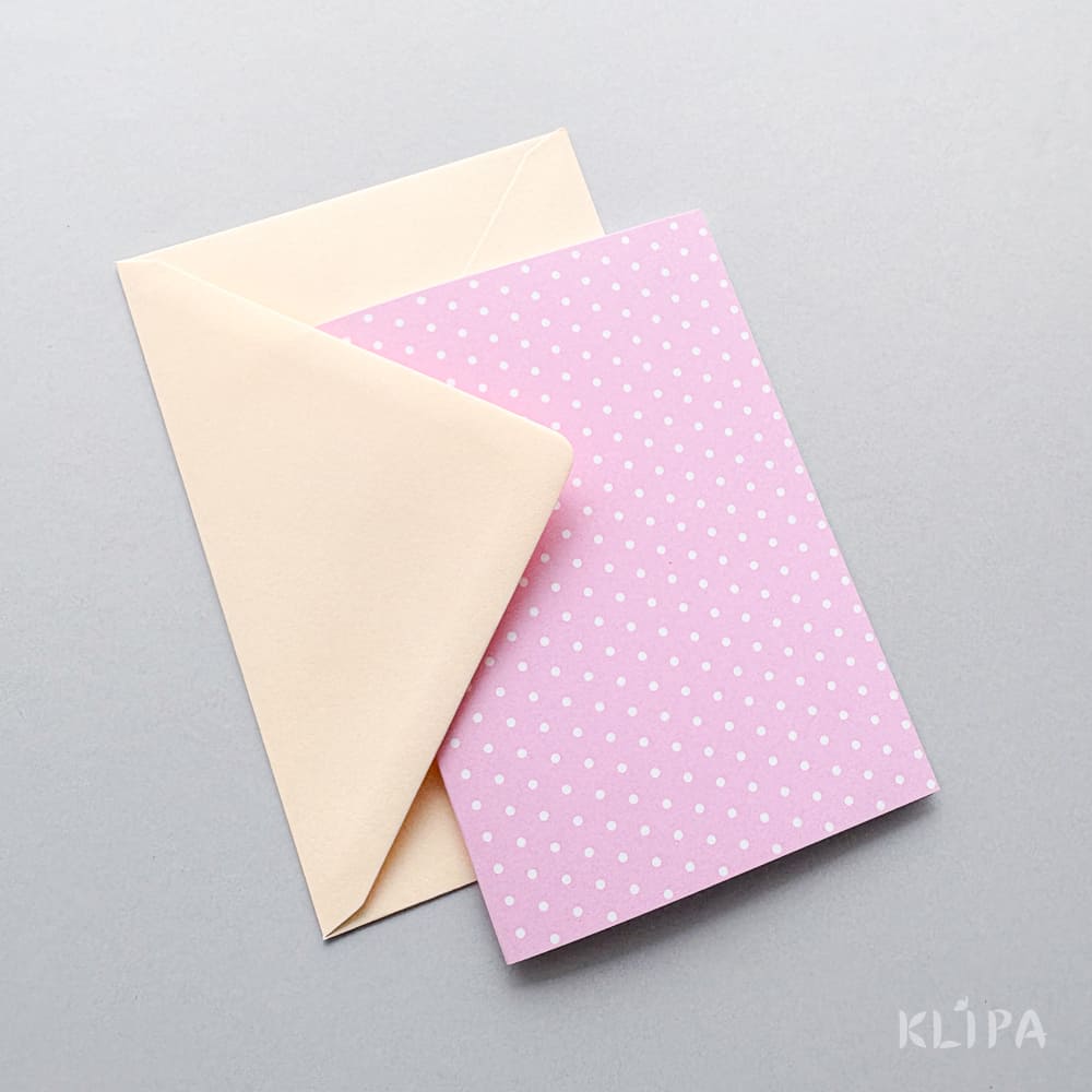 Pink greeting card with white polka dots laying over a beige envelope on a light gray surface.